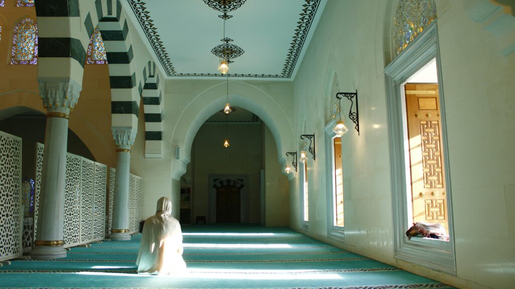 pexels-photo-31624152-31624152 A person prays in a sunlit mosque interior, showcasing Islamic architecture.