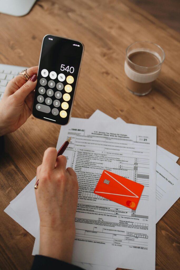 pexels-photo-6927549-6927549 A person uses a smartphone calculator over tax documents on a wooden table.