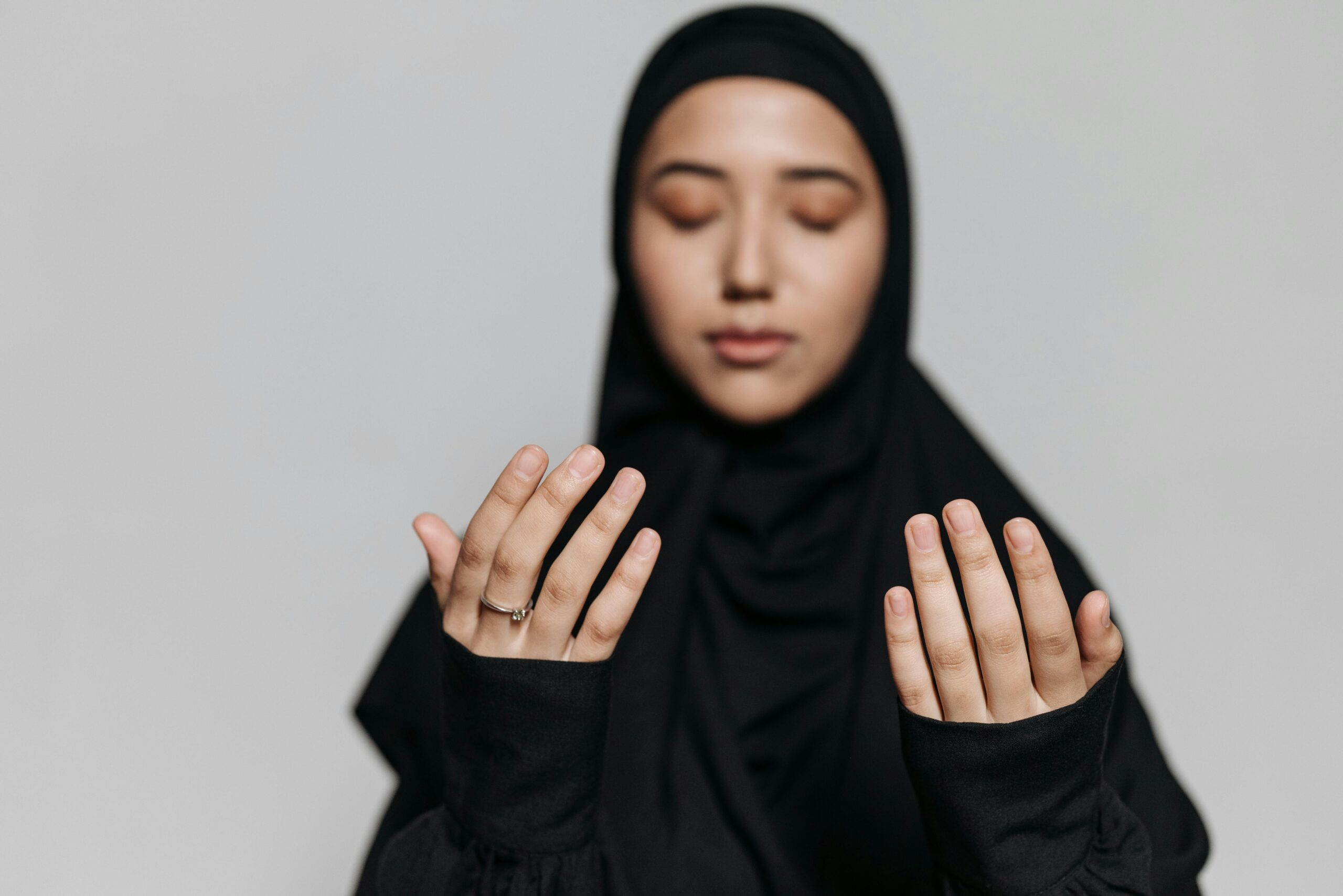 A young Muslim woman in traditional attire prays with her hands raised and eyes closed, symbolizing devotion.