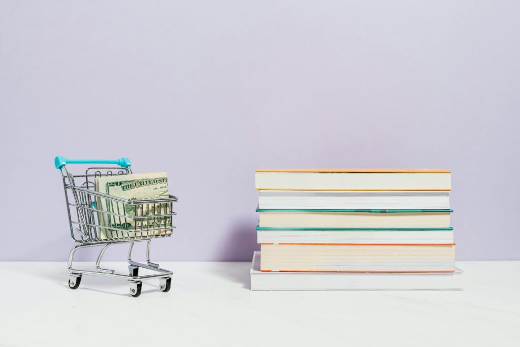 pexels-photo-5632400-5632400 A shopping cart with dollar bills next to a stack of textbooks on a light background.