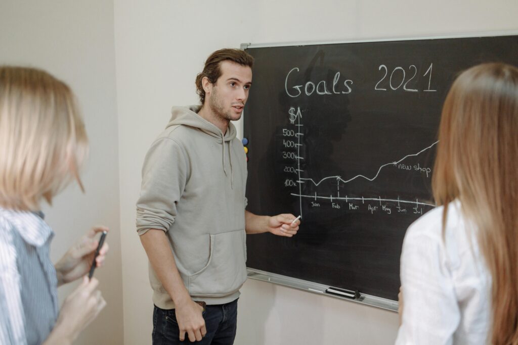 pexels-photo-5961129-5961129 Three professionals engaged in a business discussion about goals written on a chalkboard in an office.