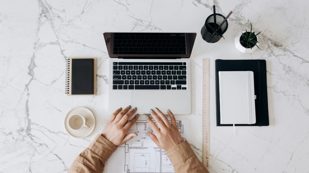 pexels-photo-6894103-6894103 Top view of a stylish home office desk with a laptop, planner, and coffee cup, showing hands on a blueprint.